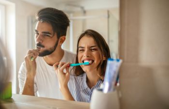 A cheerful young couple brushing their teeth in front of a bathroom mirror.